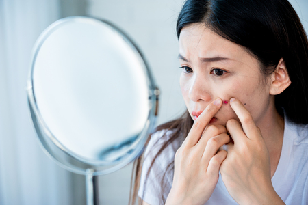 Asian teenage woman  looking at mirror and squeeze acne problem on her face, skin care concept.の写真素材