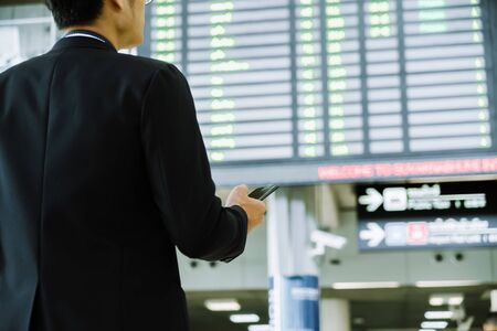 Elegant asian businessman checking email on mobile phone while walking with suitcase inside airport, experienced male employer using cell telephone while waiting for taxi car coming before work travelの写真素材