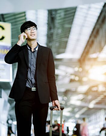 Elegant asian businessman checking email on mobile phone while walking with suitcase inside airport, experienced male employer using cell telephone while waiting for taxi car coming before work travelの写真素材