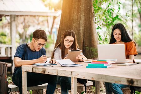 A group of young or teen Asian student in university smiling and reading the book and look at the tablet or laptop computer in summer holiday.の写真素材
