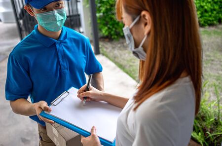 Asian deliveryman delivering package parcel food to customer in front of her house and sign to receive the box after she ordered online by application during the outbreak of the coronavirus or Covid-19 virus.の写真素材