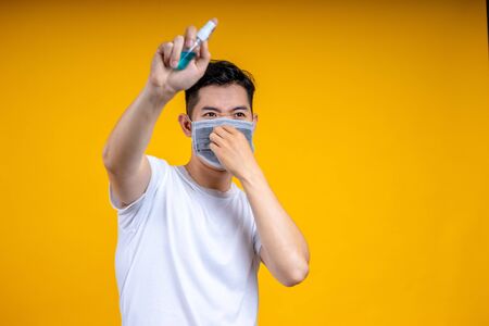 Asian man is wearing a white t-shirt and wearing a mask, holding an alcohol spray bottle. He was spraying it for cleaning  or washing his hands to prevent an outbreak of coronavirus or covid-19.の写真素材