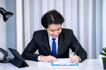 Asian business man dringking coffee wearing suit, enjoying watching educational webinar on laptop or laptop and smartphone. Smiling with chat and holding video call with clients partners.の写真素材