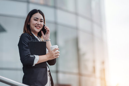 Young Asian businesswoman talking on phone and sitting in airport before business trip. Beautiful woman passenger has mobile call and discusses something with smile, holds coffee in handの写真素材