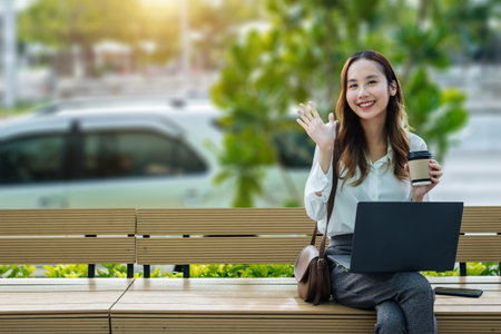 Smiling Asian curly business woman wearing trendy sitting in the central city street and uses her phone and laptop. Pretty summer woman in white shirt walks down the street looking at her mobile phoneの写真素材