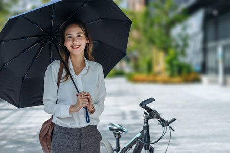 Young Asian businesswoman holding umbrella looking the watch and standing on a city street with bicycle looking at phone.の写真素材
