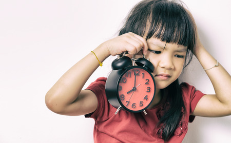 Little asian girl is angry at the alarm clock for waking her up. A kid in red shirt is upset that the alarm clock wake up her so early. Lazy Japanese girl doesn't want to get up in the morning.の写真素材