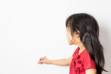 Little Asian girl is writing on a white wall copy space.の写真素材