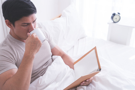 Asian male is reading book and drink coffee on morning bedの写真素材