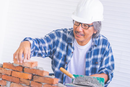 Old construction worker is laying down brick wall with cement, wearing protected builder hard hat