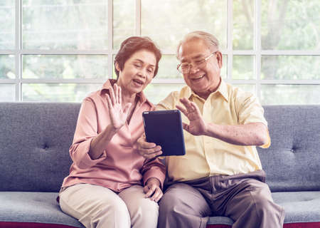 Senior couple is waving to family on Video Call communication technology on tablet on a sofa at homeの写真素材