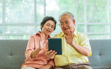 Senior couple is waving to family on Video Call communication technology on table on a sofa at homeの写真素材