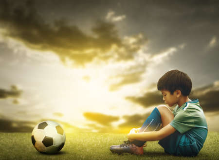 Sad soccer kid sitting on a field with soccer ball godlen sky for disappointment in Foot ball sport playing.の写真素材