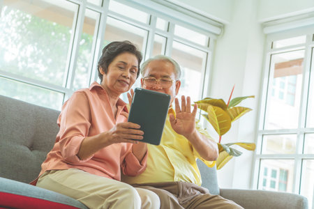 Senior couple is waving to family on Video Call communication technology on table on a sofa at home