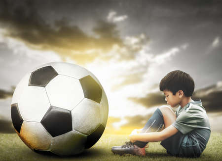 Sad soccer kid sitting on a field with Big soccer ball for obstacle in Football sport playing.の写真素材