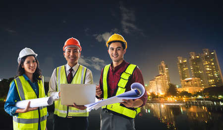 Three engineer male and female with blueprint and computer on bright Modern night Bangkok city with Furturistic technology light.の写真素材