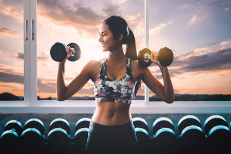 Sporty woman is holding fitness dumbbell with smile working out with sunset cloud sky in the backgroundの写真素材
