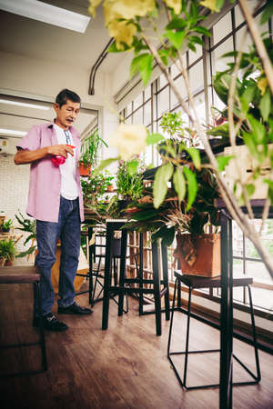 Happy Asian retirement senior is spraying water on plants and flower pot in a greenhouse for hobby at home.の写真素材