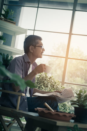 Senior Asian man is having morning coffee while reading newspaper in his houseplant garden at home.の写真素材