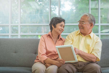 Senior Asian couple is looking at memory photo on a couch at home for happy retirement lifestyle conceptの写真素材