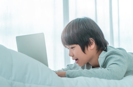 Happy Little Asian boy is using laptop computer on his bed in his room in daytime by the windows.の写真素材