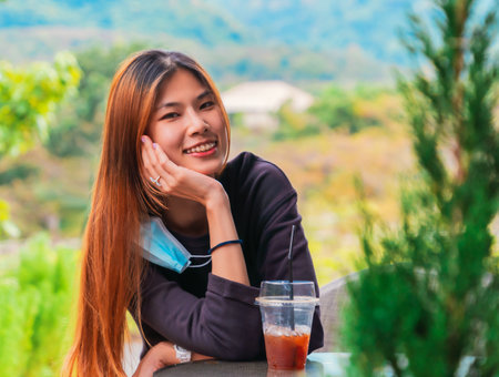 Beauty Young Asian smiling female is sitting in Cafe with forest nature background with Iced coffee on the table.の写真素材