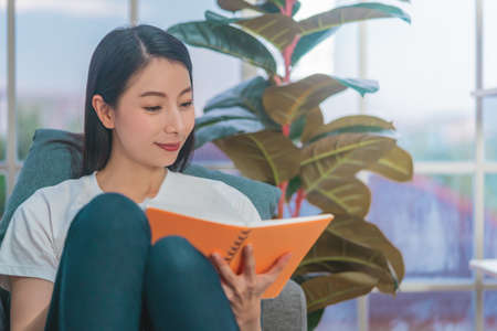 Asian woman reading orange book with smile on a sofa at homeの写真素材