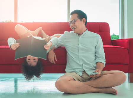 Father and son is having fun reading and studying at home on a red sofa in the livingroom. Boy is upside down while reading funny book.の写真素材