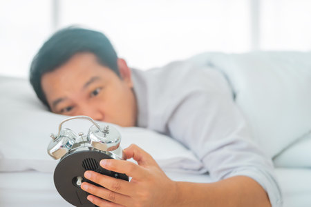 exhausted man sleeping in bed is tired by waking up to the alarm clock ring in the morning suffering...