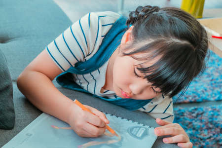 LLittle Asian girl is drawing and coloring on a book on a sofa at home for creative educational concept.の写真素材