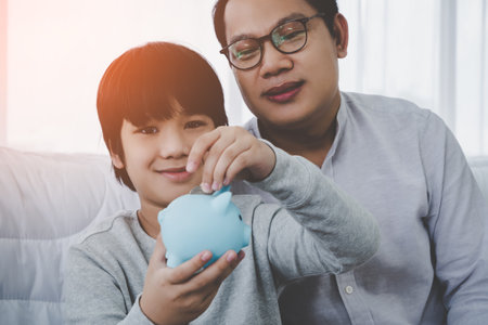Asian Father is teaching his son in money saving by inserting coin into a blue piggy bank for Financial Education concept.の写真素材