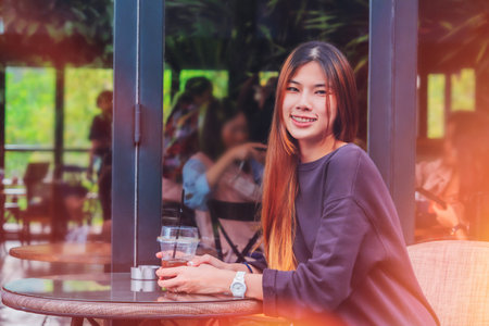 Beauty Young Asian smiling female is sitting in Cafe with forest nature background with Iced coffee on the table.の写真素材