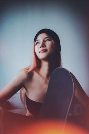 portrait of sexy Asian girl with street fashion holding a skateboard on a sofa indoor.の写真素材
