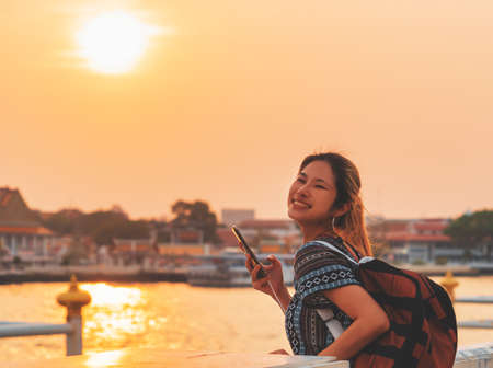 Asian woman traveler taking a photo at the Chao Phraya river pier with temple sunset reflection in the background, Bangkok, Thailand.の写真素材