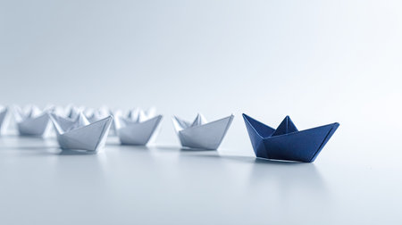 This image showcases a conceptual arrangement of paper boats. A solitary blue boat leads a sequence of white boats, set against a plain background. The minimalist aesthetic highlights the contrast in color and form. Suitable for illustrating concepts such as leadership, teamwork, or differentiation in business contexts.の素材