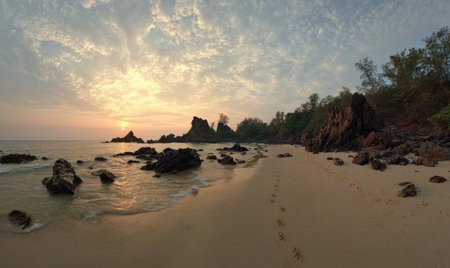 Panoramic beach scene at dawn, with rocky formations, footprints in the sand, and cloudy skyの素材