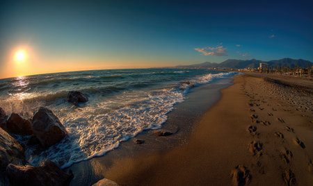 Panoramic beach view at sunset, waves crash on shore, mountains in the distanceの素材