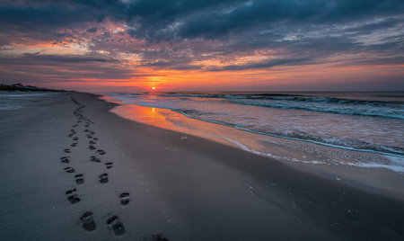 A coastal scene captures sunrise over ocean waves, footprints along the wet sandの素材