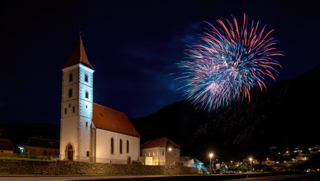 A church with a tall tower is illuminated against a dark night sky, with colorful fireworks exploding above. The scene features a small town with lights along the street. The composition highlights a celebratory event, suitable for editorial or commercial applications related to festivals and events.の素材