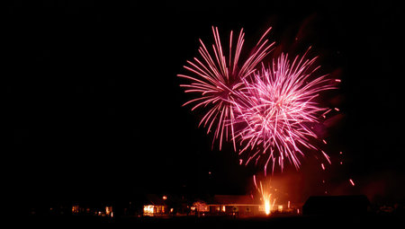 A nighttime scene features bright bursts of fireworks against a dark backdrop. The vivid pink and red explosions create a dynamic display. Below, the silhouettes of structures are subtly illuminated, adding depth to the overall composition. This image is suitable for various commercial applications, offering a visual representation of celebration and festivity.の素材