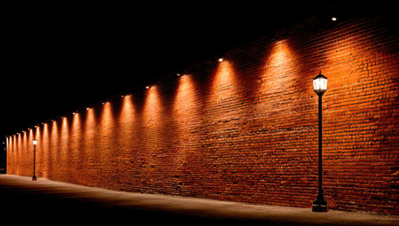 A brick wall is illuminated by a series of spotlights and street lamps, creating a contrast of light and shadow against a dark background. The texture of the brick is visible, with the warm glow suggesting a nighttime setting. This image could be used for various design projects or editorial purposes.の素材