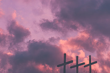The image showcases three crosses silhouetted against a vivid, cloud-filled sky. The sky features hues of pink, purple, and orange, suggesting a sunset. The composition and lighting create a dramatic and contemplative atmosphere. Suitable for illustrating themes of faith, sacrifice, and reflection, and potentially applicable to various editorial or commercial projects.の素材