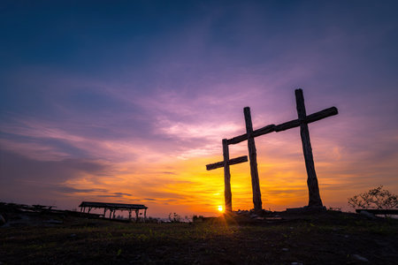 Three large wooden crosses stand silhouetted against a vibrant sunset. The image features a dramatic sky with rich colors, including orange, purple, and blue hues. The composition showcases the crosses in a landscape setting, suggesting a time of day. This image could be used for editorial or commercial purposes.の素材