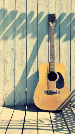An acoustic guitar stands against a weathered wooden wall, casting a long shadow. The instrument's warm tones contrast with the cool, muted color of the wall. Sunlight illuminates the scene, creating a rustic aesthetic. This image could be used for various commercial purposes including music and lifestyle content.の素材