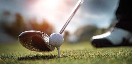 A close-up shot of a golf club about to strike a golf ball resting on a tee, set against a backdrop of green grass and soft-focus foliage. The composition utilizes overhead lighting, creating highlights and shadows. This image could be used in advertising materials or illustrations related to sports and recreation.の素材