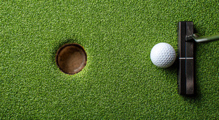 A golf ball sits near a hole with a putter in a close-up shot. The scene features a textured, green artificial turf surface. The composition uses a simple overhead perspective. This image is suitable for various uses including marketing and educational materials.の素材