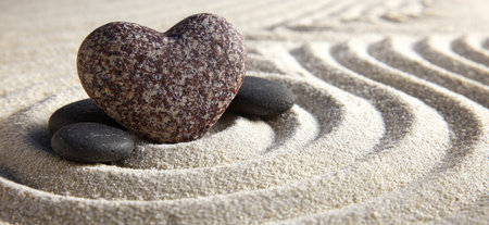 A heart-shaped stone rests atop dark stones on a textured sand surface. The sand is raked with gentle curved lines, creating a serene aesthetic. The composition presents a sense of calm with warm, neutral tones. Suitable for design projects, wellness concepts, or editorial purposes.の素材