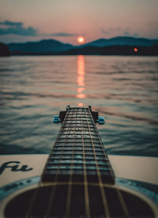 An acoustic guitar, viewed from the neck down, is centered with the lake as the backdrop, reflecting the sunset. The image shows calm water, silhouetted mountains, and a warm color palette. This tranquil scene would be suitable for use in editorial content or creative marketing materials.の素材