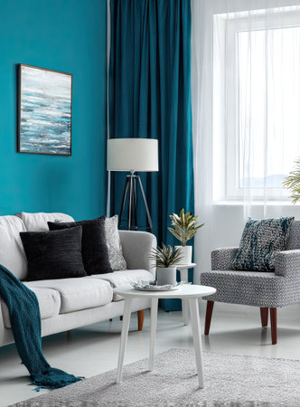 A contemporary interior presents a living room with a teal wall, white sofa, and a gray armchair. The scene features dark curtains, a framed picture, and a small table. Natural sunlight streams through a window, illuminating the room, which may be suitable for lifestyle or design content.の素材