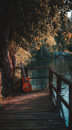 An acoustic guitar rests against a tree on a wooden dock beside a tranquil body of water. The scene displays warm tones, with sunlight filtering through foliage. The composition includes natural elements, creating a peaceful outdoor setting suitable for various creative projects and editorial use.の素材